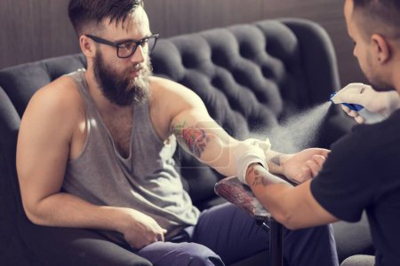 Téléchargez les photos : Tatoueur masculin tenant un pistolet de tatouage, montrant un processus de fabrication de tatouages sur le bras d'un mannequin tatoué masculin. - en image libre de droit