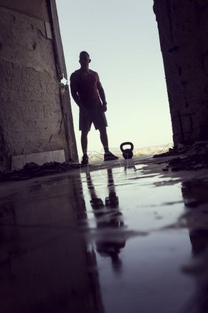 Téléchargez les photos : Reflet d'un jeune athlète musclé et athlétique qui s'entraîne avec une kettlebell dans un bâtiment en ruine à côté d'une flaque d'eau - en image libre de droit