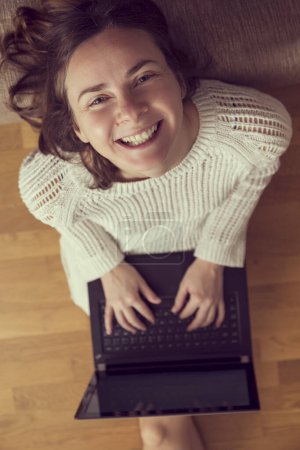 Foto de Vista superior de una joven sentada en el suelo de la sala de estar, apoyada en un sofá y usando una computadora portátil - Imagen libre de derechos