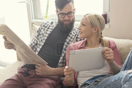 Photo for Young couple in love sitting on a couch in their apartment, enjoying their free time, reading newspaper and surfing the web on a tablet computer - Royalty Free Image