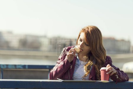 Téléchargez les photos : Jeune belle brune debout dans un escalier, buvant du café, mangeant un cookie, surfant sur Internet sur une tablette et profitant d'une journée ensoleillée - en image libre de droit