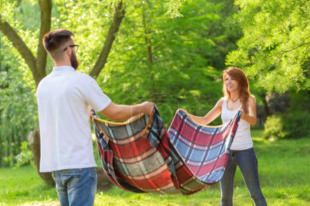 Téléchargez les photos : Couple amoureux plaçant une couverture de pique-nique sur une prairie. Loisirs et détente dans la nature - en image libre de droit