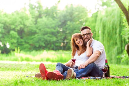 Photo for Couple in love sitting on a picnic blanket in a park holding glasses of wine, hugging and enjoying their time together - Royalty Free Image