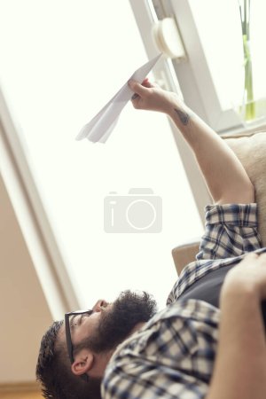 Téléchargez les photos : Un beau mec allongé sur un canapé près de la fenêtre, tenant un avion en papier et rêvant. Effet fusée éclairante sur la fenêtre - en image libre de droit