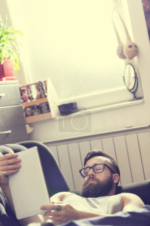 Téléchargez les photos : Modèle masculin couché sur un canapé dans un salon, surfant sur le web sur une tablette - en image libre de droit