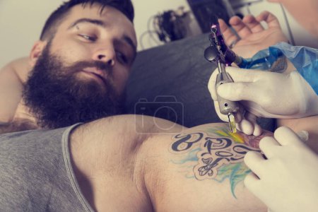 Téléchargez les photos : Tatoueur masculin tenant un pistolet de tatouage, montrant un processus de fabrication de tatouages sur le bras d'un mannequin tatoué masculin. Se concentrer sur le tatouage d'un modèle - en image libre de droit