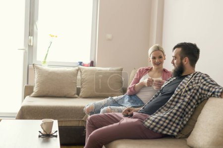 Téléchargez les photos : Couple amoureux profiter de leur temps libre, assis sur un canapé à côté de la fenêtre, boire du café et bavarder. Effet fusée éclairante sur la fenêtre - en image libre de droit