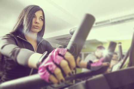 Photo for Two young people working out in a gym, riding a bike and working out on a ski simulator. Focus on the girl, lens flare effect - Royalty Free Image