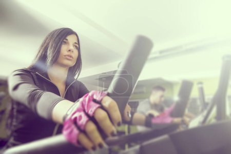 Photo for Two young people working out in a gym, riding a bike and working out on a ski simulator. Focus on the girl, lens flare effect - Royalty Free Image