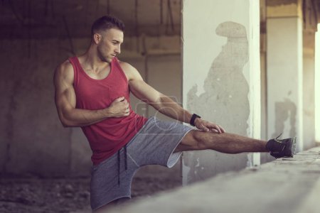 Téléchargez les photos : Musclé, athlétique, jeune homme levant sa jambe sur une partie d'un mur d'un bâtiment abandonné en ruine qui s'étire avant l'entraînement - en image libre de droit