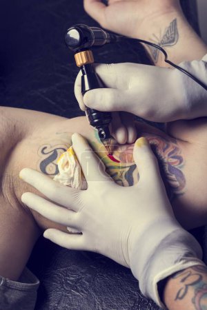 Photo for Male tattoo artist holding a tattoo gun, showing a process of making tattoos on a male tattooed model's arm. - Royalty Free Image