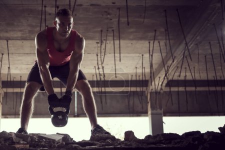 Photo for Front view of a young muscular athlete working out, lifting a kettlebell weight in an abandoned ruined building - Royalty Free Image