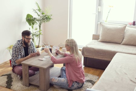 Téléchargez les photos : Jeune couple amoureux assis par terre à côté de la table, jouant aux cartes, buvant du jus et s'amusant. Effet fusée éclairante sur la fenêtre - en image libre de droit