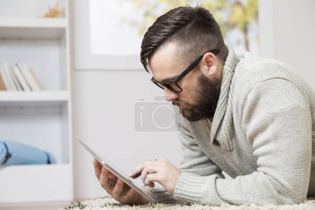 Téléchargez les photos : Homme couché sur le sol, tenant une tablette et lisant un livre électronique - en image libre de droit