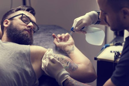 Téléchargez les photos : Tatoueur masculin tenant un pistolet de tatouage, montrant un processus de fabrication de tatouages sur le bras d'un mannequin tatoué masculin. - en image libre de droit