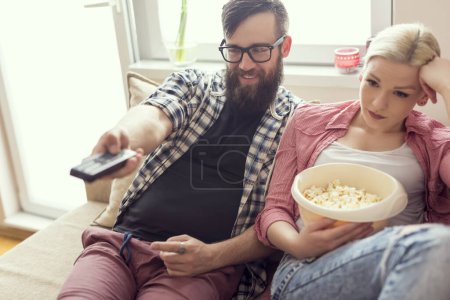 Téléchargez les photos : Les jeunes couples se détendent dans leur appartement, allongés sur le canapé, regardant un film et mangeant du pop-corn. Concentrez-vous sur le gars, effet de fusée éclairante sur la fenêtre - en image libre de droit