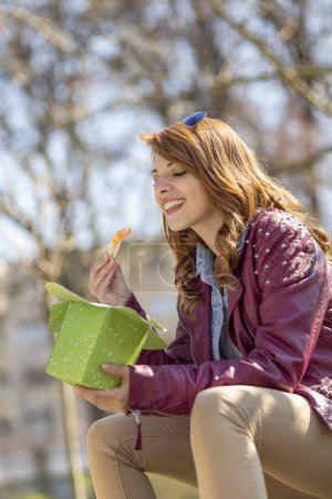 Téléchargez les photos : Jeune, belle brune assise sur un banc en bois dans un parc par une journée ensoleillée à manger de la nourriture chinoise - en image libre de droit