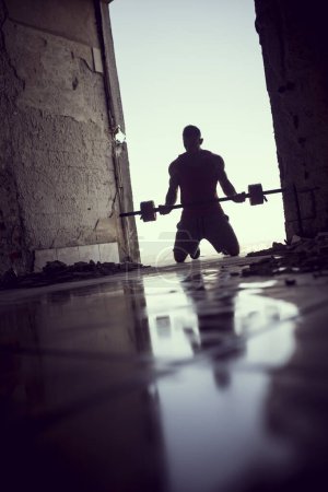 Téléchargez les photos : Musclé, athlétique, jeune athlète soulevant des poids dans un bâtiment en ruine à côté d'une flaque d'eau - en image libre de droit