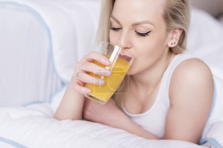 Foto de Hermosa mujer joven acostada y bebiendo un jugo de naranja recién hecho en la cama - Imagen libre de derechos