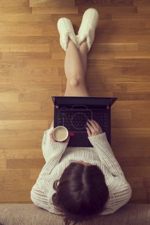 Foto de Vista superior de una joven sentada en el suelo de la sala de estar, apoyada en un sofá, tomando café y usando una computadora portátil - Imagen libre de derechos