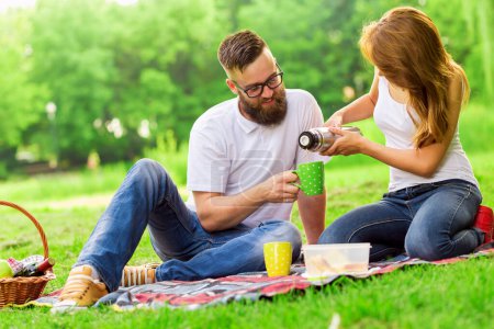 Foto de Pareja joven en un picnic en un parque. Chica vertiendo café caliente de una botella de termo. Concéntrate en la botella de taza y termo - Imagen libre de derechos