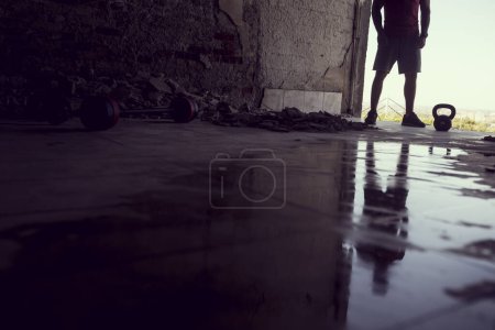 Photo for Reflection of a muscular, athletic built, young athlete working out with a kettlebell in a ruin building next to a puddle of water - Royalty Free Image