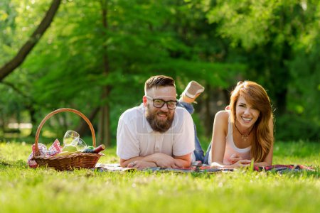 Téléchargez les photos : Couple couché sur une couverture de pique-nique dans un parc avec un panier de pique-nique rempli de fruits, de nourriture et de vin placé à côté d'eux - en image libre de droit