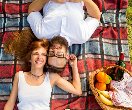 Photo for Top view of couple in love lying on a picnic blanket in the park with basket full of food and drinks placed next to them - Royalty Free Image