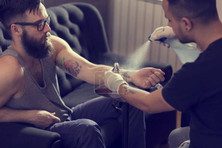 Foto de Artista de tatuajes masculino sosteniendo una pistola de tatuaje, mostrando un proceso de hacer tatuajes en el brazo de un modelo tatuado masculino. - Imagen libre de derechos
