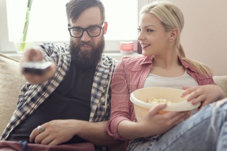 Téléchargez les photos : Les jeunes couples se détendent dans leur appartement, allongés sur le canapé, regardant un film et mangeant du pop-corn. Effet fusée éclairante sur la fenêtre, se concentrer sur le gars - en image libre de droit