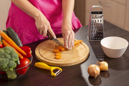 Téléchargez les photos : Détail des mains féminines coupant des carottes fraîches avec un couteau de cuisine sur une planche à découper pour faire de la salade de légumes frais - en image libre de droit