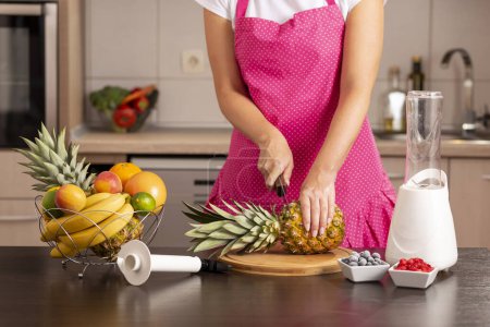 Woman cutting a pineapple top with a kitchen knife on a cutting board in order to peel it with a pineapple cutter