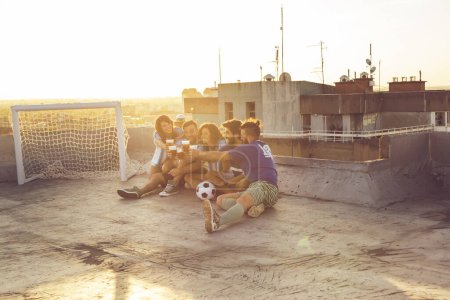 Foto de Grupo de jóvenes sentados en la azotea de un edificio, con camisetas, descansando después de un partido de fútbol, bebiendo cerveza y haciendo un brindis - Imagen libre de derechos