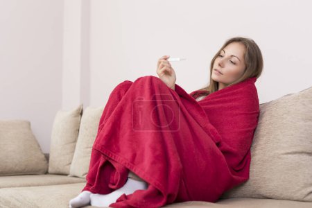 Photo for Young woman sitting on a couch covered with blanket, having a fever, holding a thermometer after measuring temperature. - Royalty Free Image