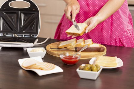 Téléchargez les photos : Détail des mains féminines étalant du beurre sur une tranche de pain ; femme faisant des sandwichs chauds dans une machine à sandwich pour le petit déjeuner. Focus sur le couteau - en image libre de droit
