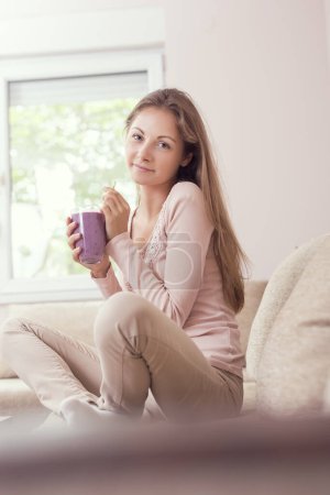 Photo for Beautiful girl sitting on a living room couch, smiling and holding a glass of raspberry and blueberry mix smoothie - Royalty Free Image