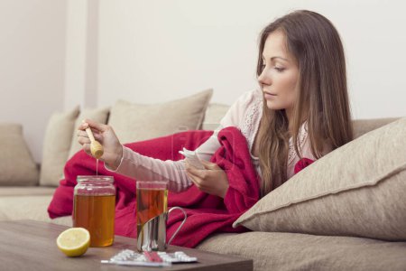 Foto de Mujer enferma acostada en la cama con fiebre alta y gripe, poniendo un poco de miel en su té. Limón, tarro de miel y pastillas alrededor de la mesa - Imagen libre de derechos
