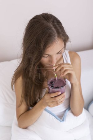 Photo for Beautiful girl sitting on a bed after waking up in the morning and drinking a raspberry and blueberry mix smoothie - Royalty Free Image