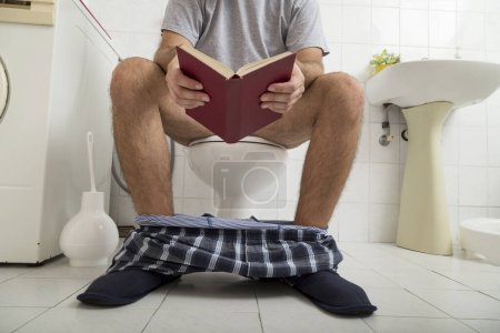 Téléchargez les photos : Détail d'un homme assis sur les toilettes avec son pyjama et ses sous-vêtements, lisant un livre - en image libre de droit