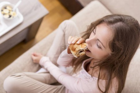 Foto de Hermosa chica sentada en un sofá de la sala de estar, comiendo un croissant para el desayuno. Enfoque selectivo - Imagen libre de derechos