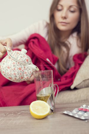 Téléchargez les photos : Femme malade couchée au lit avec une forte fièvre et une grippe, versant du thé d'une théière dans un verre. Citron et pilules sur la table, se concentrer sur la théière - en image libre de droit