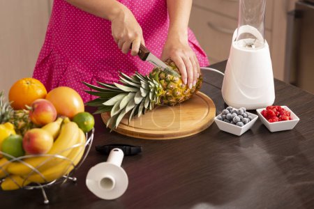 Téléchargez les photos : Vue en angle élevé d'une femme coupant un dessus d'ananas avec un couteau de cuisine sur une planche à découper afin de le peler avec un coupe-ananas - en image libre de droit