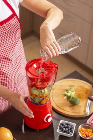 Vista de ángulo alto de las manos femeninas vertiendo agua en un tazón de licuadora sobre verduras frescas para hacer caldo de verduras. Concéntrate en el agua