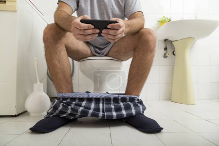 Téléchargez les photos : Détail d'un homme assis sur les toilettes, lisant le journal sur un téléphone intelligent - en image libre de droit
