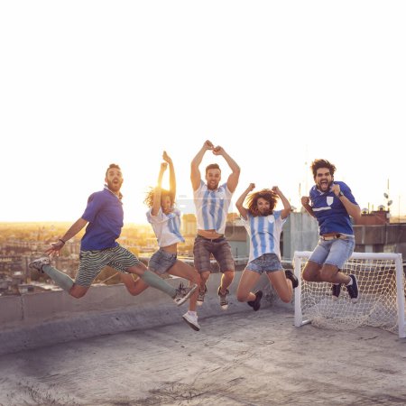 Téléchargez les photos : Groupe de jeunes amis s'amusant sur un toit de bâtiment après un match de football ; sauter, tous pris dans l'air avec le paysage urbain et le coucher du soleil en arrière-plan - en image libre de droit