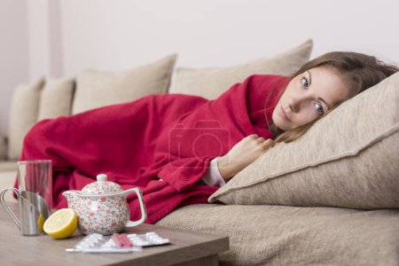 Foto de Mujer enferma cubierta con una manta acostada en la cama con fiebre alta y gripe. Pastillas, tetera y limón sobre la mesa - Imagen libre de derechos