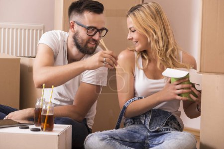 Téléchargez les photos : Couple amoureux assis sur le sol dans leur nouvel appartement entouré de boîtes en carton, manger à emporter de la nourriture chinoise et boire du thé glacé - en image libre de droit
