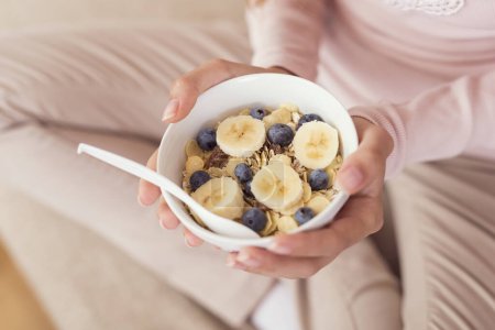 Photo for Detail of a young woman sitting on a living room couch, holding a bowl of cereal and having breakfast - Royalty Free Image