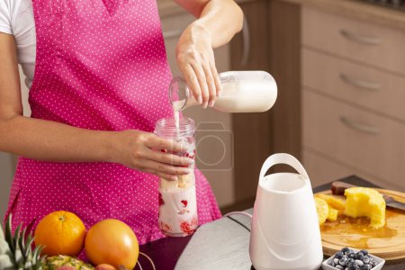 Foto de Detalle de una mujer vertiendo leche sobre las frutas en un tazón de licuadora y haciendo frambuesa fresca, plátano, leche y batido de miel para el desayuno. Concéntrate en la leche - Imagen libre de derechos