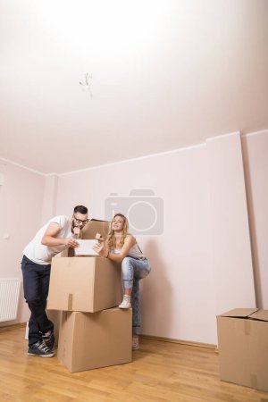 Photo for Young couple in love moving in a new apartment, standing next to cardboard boxes, holding tablet computer and surfing the web while planning to redecorate their new home - Royalty Free Image